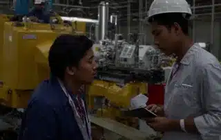 a healthcare professional consults with a factory worker next to industrial machinery.